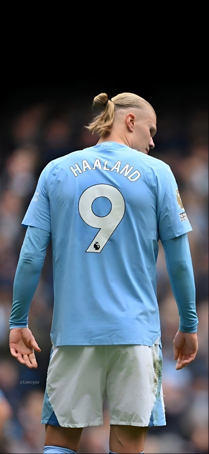 a soccer player with his hair in a ponytail and wearing a blue jersey is looking down at the ground