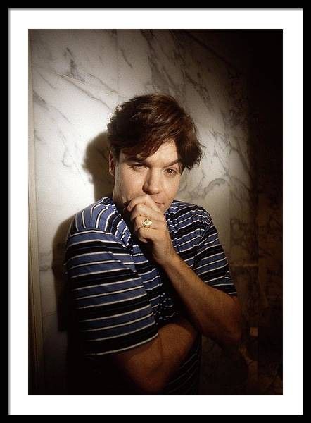 a man with his hand on his chin looking at the camera while standing in front of a wall