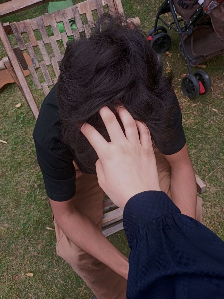 a man sitting on top of a wooden chair holding his hand to his head while looking down at the ground