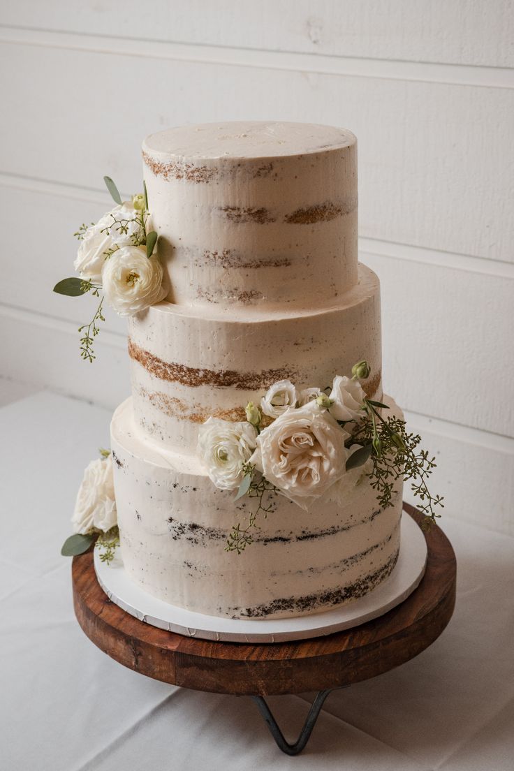 a three tiered cake with white flowers and greenery sits on a wooden platter