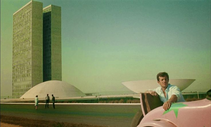 a man sitting on the hood of a pink car in front of a tall building