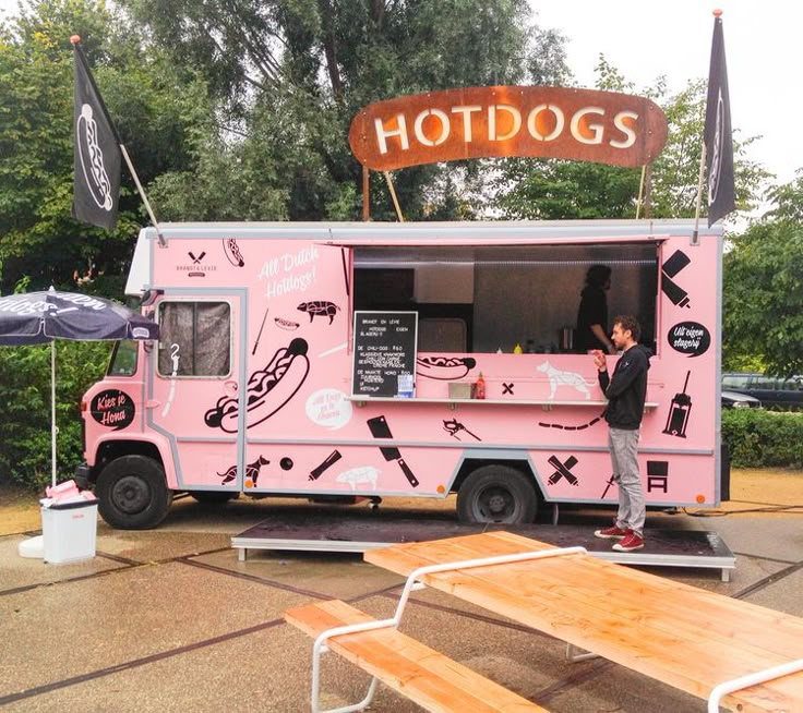 a man standing in front of a pink food truck with hot dogs written on it