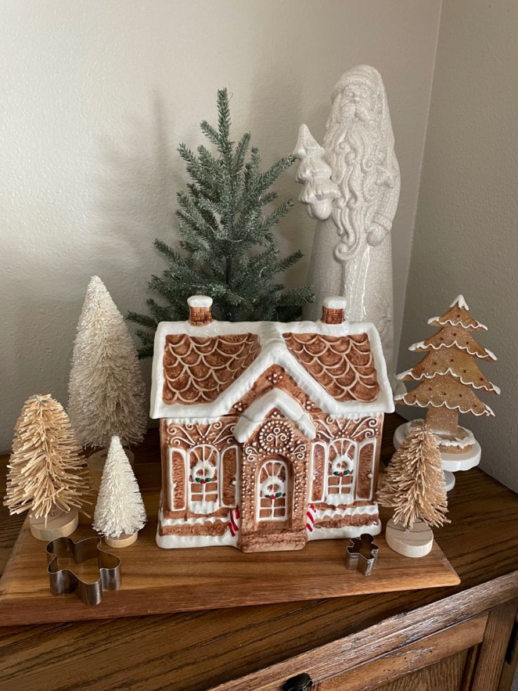 a gingerbread house with trees and other decorations on a wooden table in front of a white wall