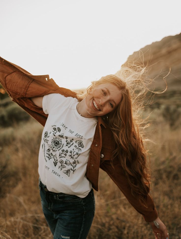 a woman with long hair is standing in a field smiling and holding her arms out to the side