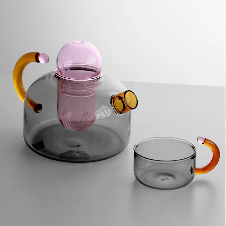 a glass teapot with a pink handle and yellow handles on the top, sitting next to a clear glass cup