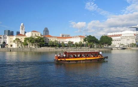 a yellow boat traveling down a river next to tall buildings in the background with people on it