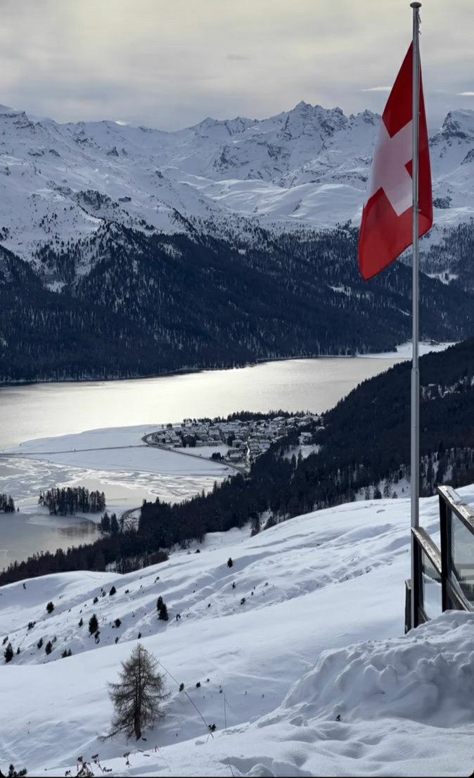 a flag on top of a snow covered hill next to a body of water with mountains in the background