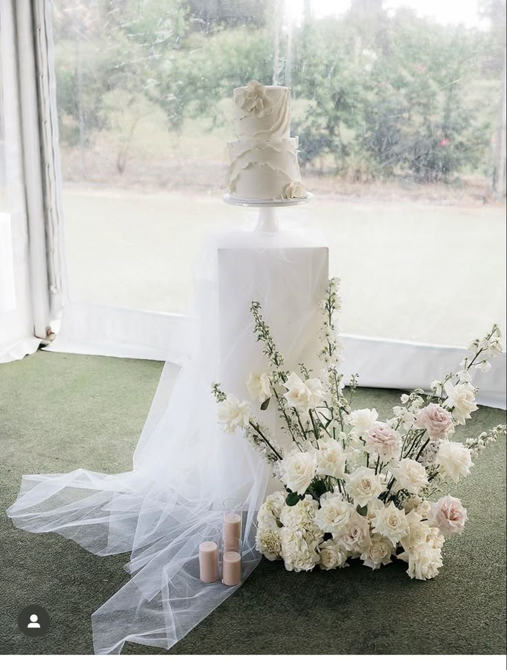 a wedding cake and flowers on the floor in front of a window with sheer curtains