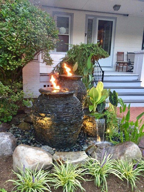 an outdoor fountain in front of a house with rocks and plants around it, surrounded by greenery