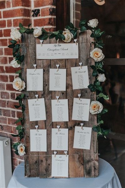 a wooden frame with seating cards attached to it and flowers on the side, along with greenery