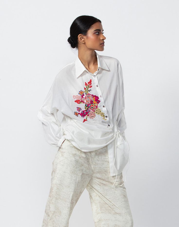 Editor's Note A batwing oversized side tie-up shirt with intricate mirror and thread hand-embroidered motif. Note: Pant worn by model is for styling purposes only Color: White Fabric: Cambric (Cotton) Components: Shirt Occasion: Daywear Care: Dry Clean Only About the Designer Saaksha & Kinni is our vision, our expression and our ode to tradition. All the while keeping on mind our tradition and roots in India – Our bold prints, metal, and stone embroidery techniques and our love of Indian fabrics White Casual Shirt With Embroidered Graphics, Oversized Embroidered Shirt For Summer, Oversized Floral Embroidery Blouse For Spring, Summer Oversized Embroidered Shirt, Oversized Floral Embroidered Blouse For Spring, Oversized Spring Blouse With Floral Embroidery, Embroidered Oversized Summer Shirt, Oversized Embroidered Summer Blouse, White Cotton Shirt With Embroidered Text