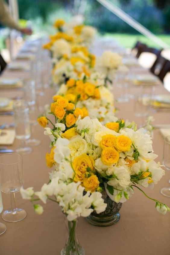 a long table with yellow and white flowers in vases on top of the tables