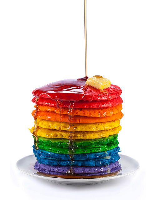 a stack of pancakes covered in rainbow icing with a wooden skewer sticking out of the top