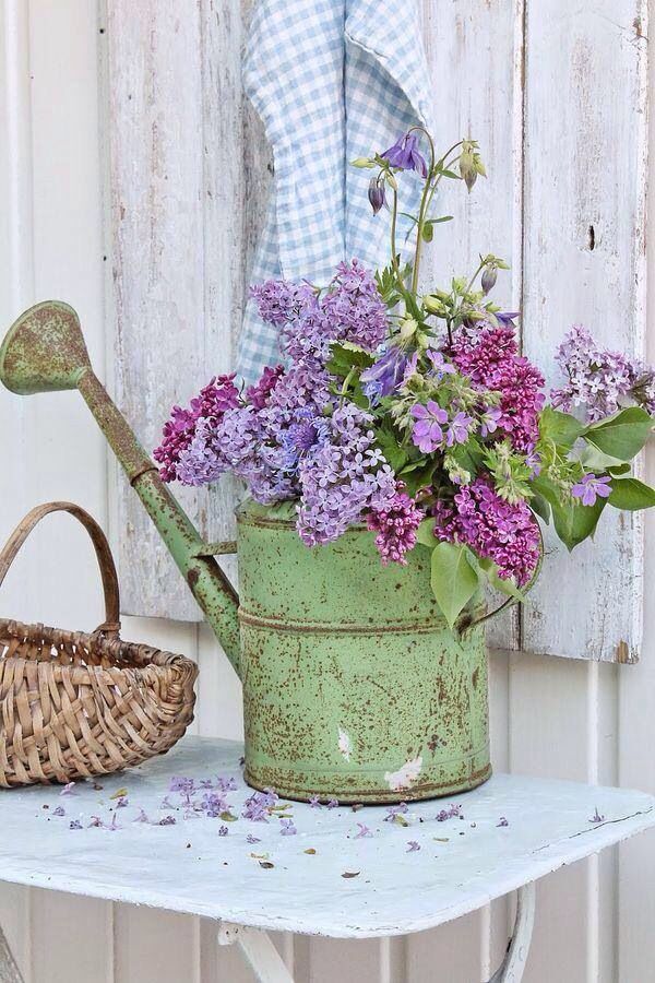 a green watering can with purple flowers in it next to a wicker basket and blue gingham towel