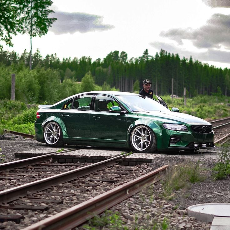 a green car parked on the side of train tracks next to a man in a black jacket