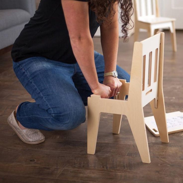 a woman kneeling down on the floor to assemble a wooden chair with one leg up