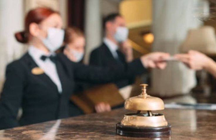 two hotel staffs wearing face masks behind a desk with a bell on it and one woman in the foreground