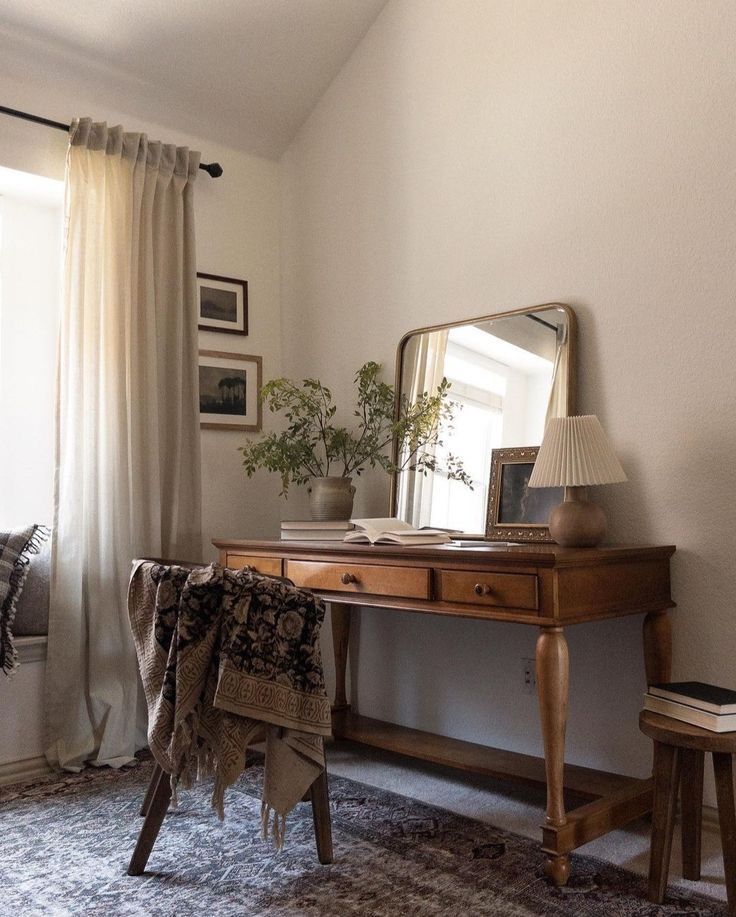 a desk with a mirror, lamp and chair in a room that has curtains on the windowsill