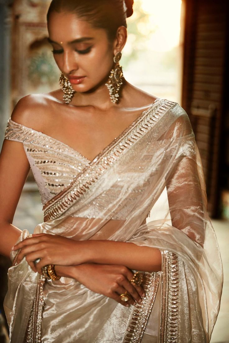 Editor's Note The Iconic Winged Blouse With Suchi Dori, Swarovski Crystals & Mini Mirrors. Silver Wrinkled Tissue Saree With Crystal Tassels & A Contemporary Hand Embroidered Border. Possibly The Most Iconic Piece From Tales Of Masai. Handwoven In Banaras, And Taken Form In The Matsya Ateliers. Color: Ivory Fabric: Tissue Components: Sari & Blouse Care: Dry Clean Only About the Designer Matsya by Utkarsh Ahuja curated for the princess in disguise, royal humility at the heart of a simple life liv Wrinkled Tissue Saree, Ivory Designer Saree, Ivory Blouse Saree, Ivory Tissue Saree, Tissue Fabric Saree, Tissue Saree Styling, Silver Tissue Saree Blouse Designs, Silver Saree Blouse Design, Tissue Lengha