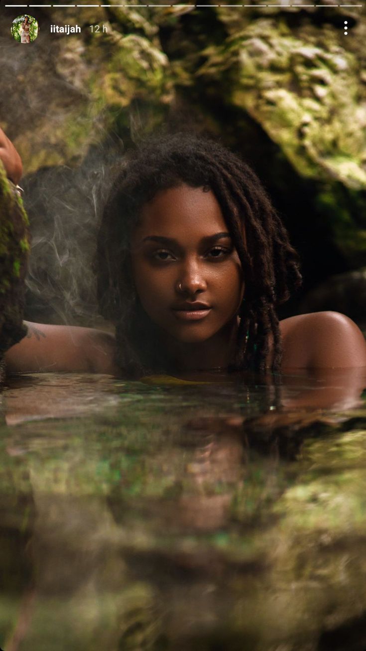 a woman with dreadlocks is in the water near rocks and mossy trees