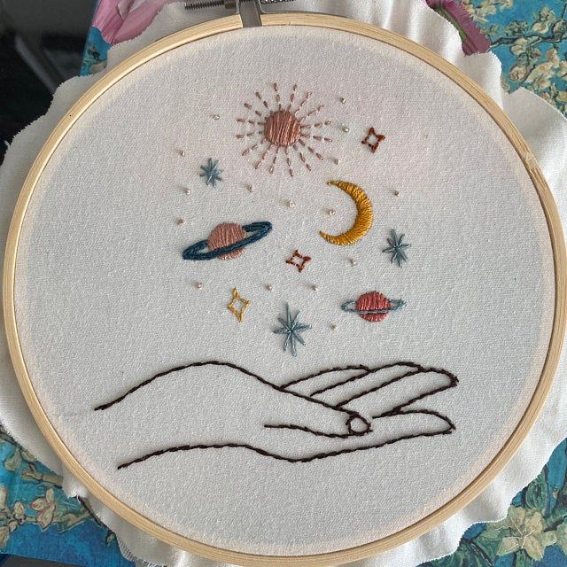 a hand is holding an object in the middle of a embroidery hoop with stars and planets on it