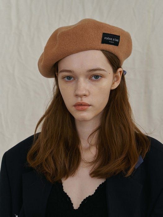 a woman with red hair wearing a brown beret and black shirt, standing in front of a white background