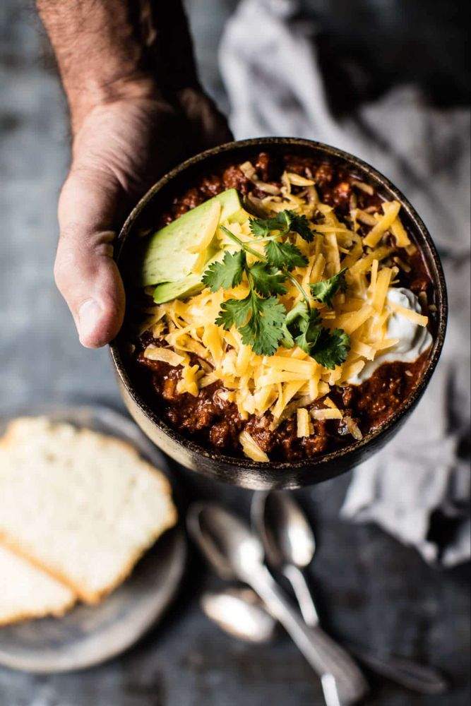 a person holding a bowl of chili with cheese and avocado on the side