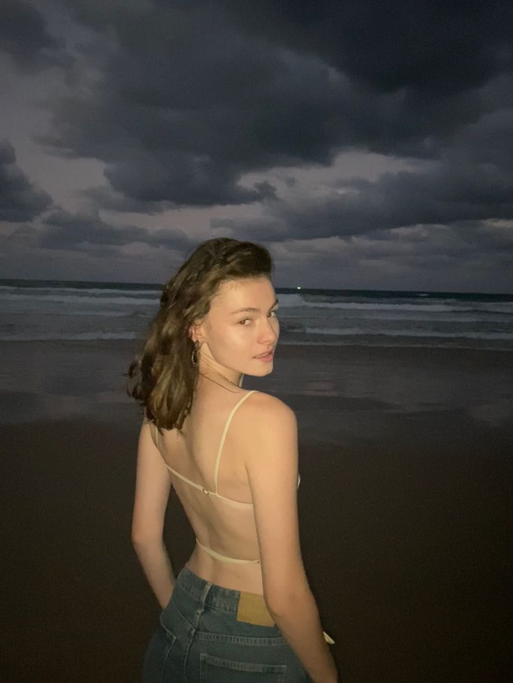 a woman standing on top of a beach next to the ocean under a cloudy sky