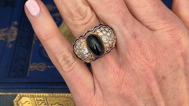 This stunning ring is centered with one (1) cabochon cut sapphire that is bezel set. On either side of center stone are fifteen (15) round diamonds that are bezel/prong set. The ring is accented with blue enamel, some enamel is damaged or missing. The ring measures 17.4mm at the top, rises 12.2mm above the finger, tapering to 2.1mm wide and 1.0mm thick at the base of the shank. It is currently a size 10. Please call with questions about the limited sizing of this ring. There is light wear to the enamel. Luxury Diamond Cabochons For Wedding, Luxury Multi-stone Cabochons For Formal Occasions, Luxury Diamond Cabochons For Formal Occasions, Luxury Hallmarked Oval Cabochon Sapphire Ring, Luxury Round Cabochons With Bezel Setting, Luxury Gold Diamond Cabochons, Luxury Cabochons With Bezel Setting, Formal Enamel Ring With Gemstone, Luxury Domed Cabochons For Formal Occasions