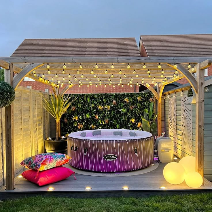 an outdoor hot tub surrounded by lights and pillows on the ground in front of a wooden pergoline