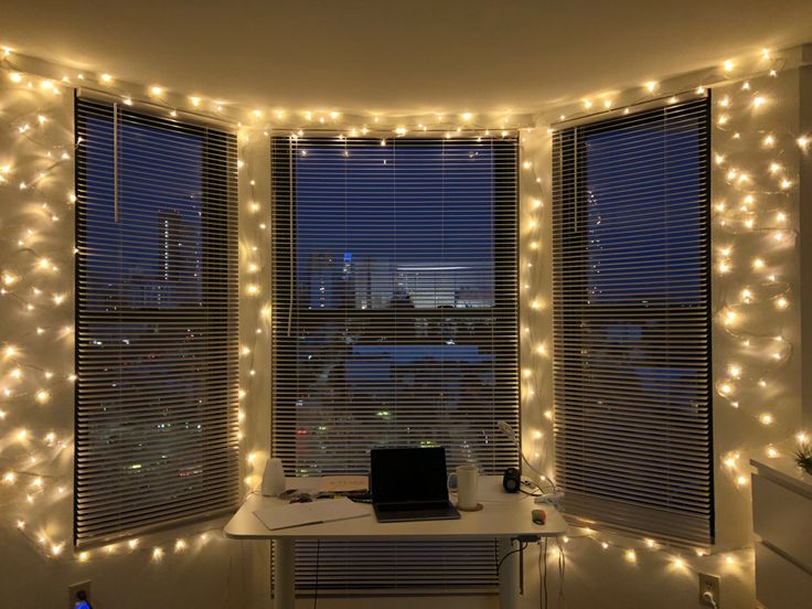 a laptop computer sitting on top of a desk in front of two windows covered with fairy lights