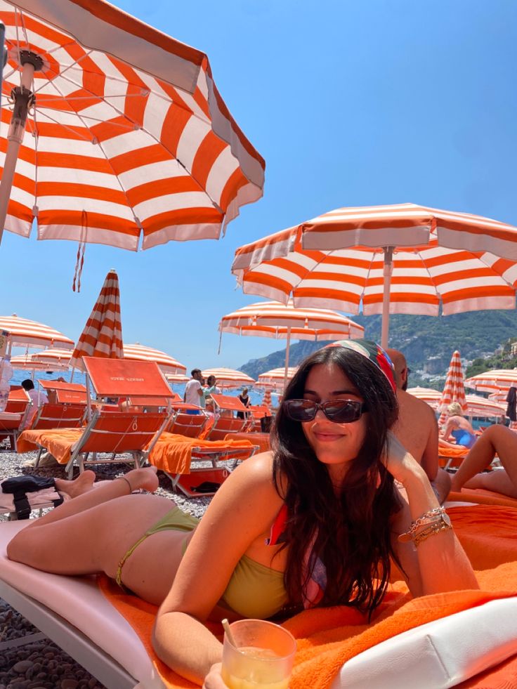 a woman laying on an orange and white beach towel next to the ocean with umbrellas