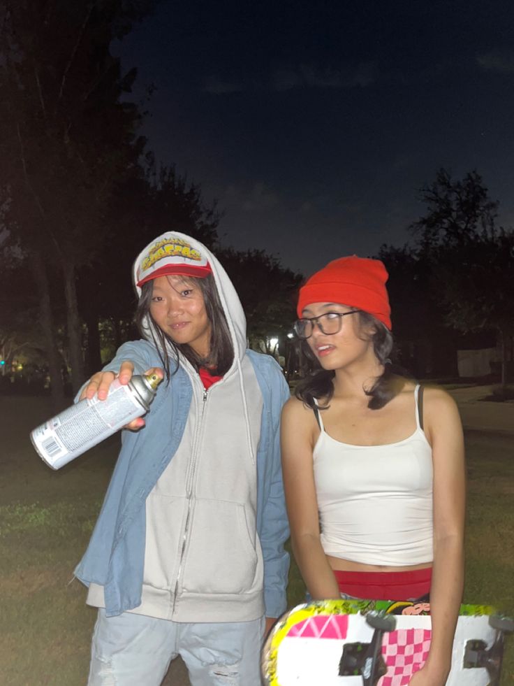 two people standing next to each other in front of a building at night, one holding a bottle and the other wearing a red hat