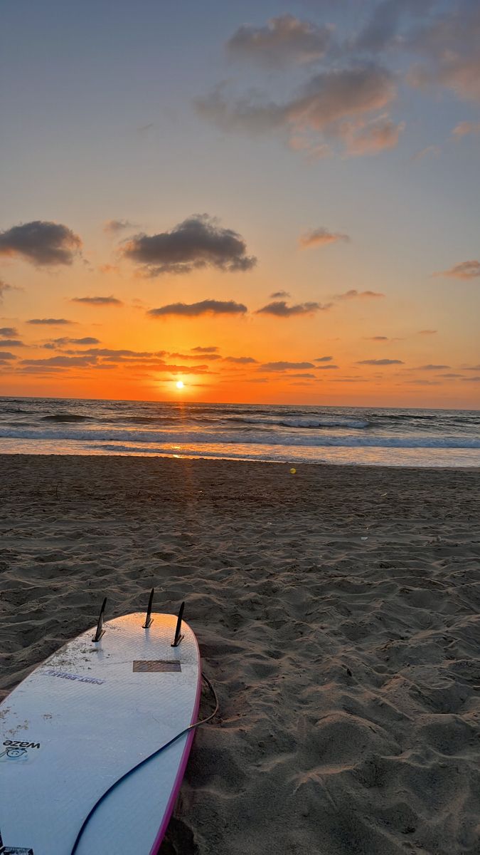 a surfboard sitting on top of a sandy beach near the ocean at sunset or dawn