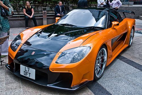 an orange and black sports car is parked on the street with people standing around it