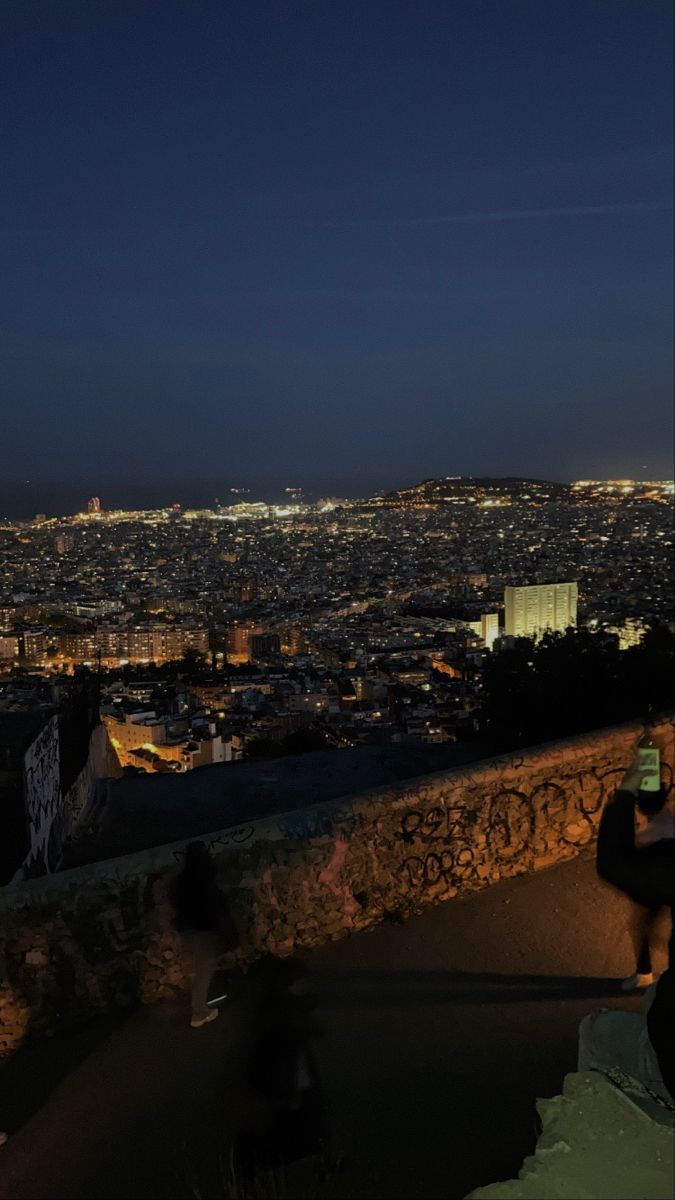 the city lights are lit up in the night sky as people sit on steps and watch