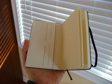 a hand holding an open notebook in front of a window with blinds on the outside