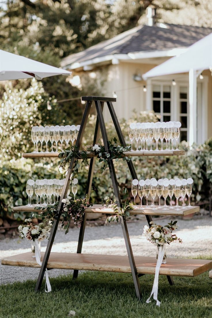 a wooden stand with wine glasses and flowers on it in front of a white house