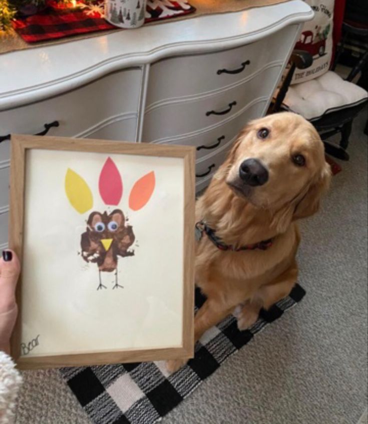 a dog sitting next to a person holding up a thanksgiving turkey painting in front of a dresser