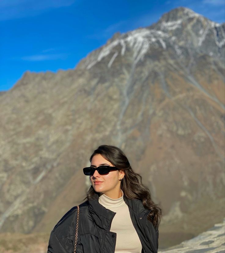 a woman wearing sunglasses standing in front of a mountain with her hand on her hip