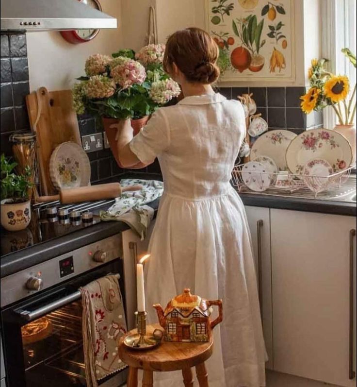 a woman in a white dress is holding flowers and looking at dishes on the stove