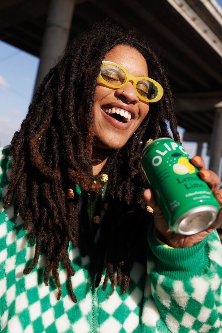 a woman with dreadlocks and glasses holding a can of green beer in front of her face