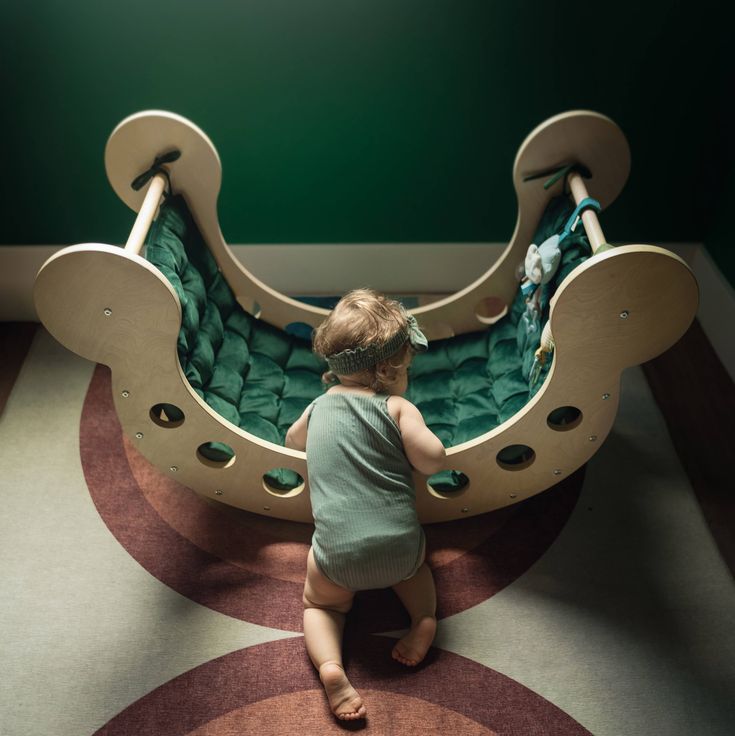 a toddler sitting on the floor in front of a wooden rocking chair with green cushions