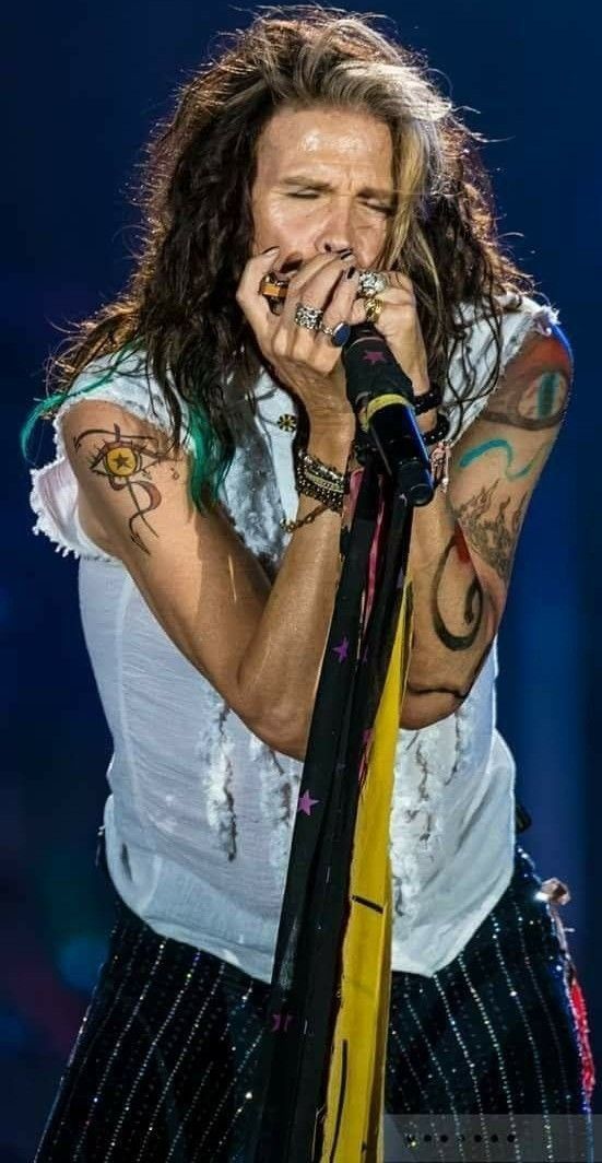 a man with long hair and piercings on his arm holding a microphone to his mouth