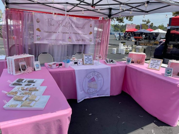 a pink table topped with lots of desserts under a white awning next to a parking lot