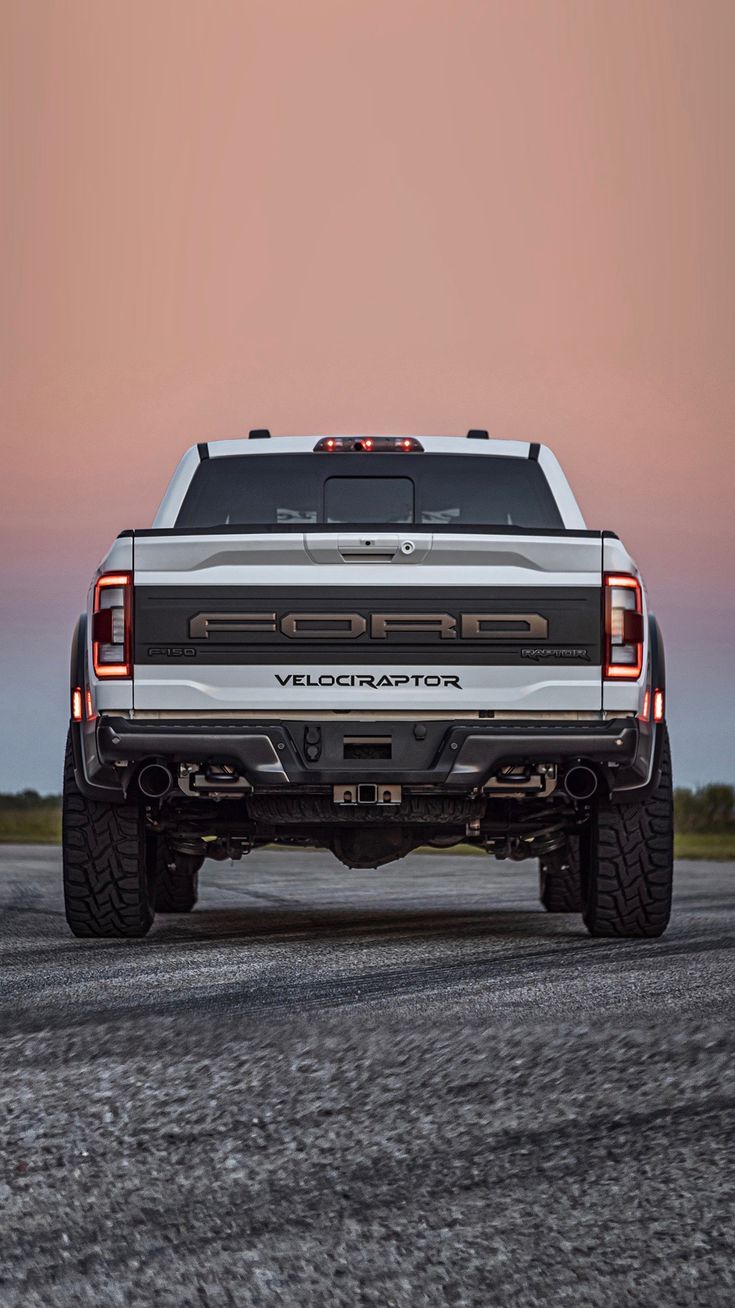 the back end of a white pickup truck on a gravel road at dusk with an orange sky in the background