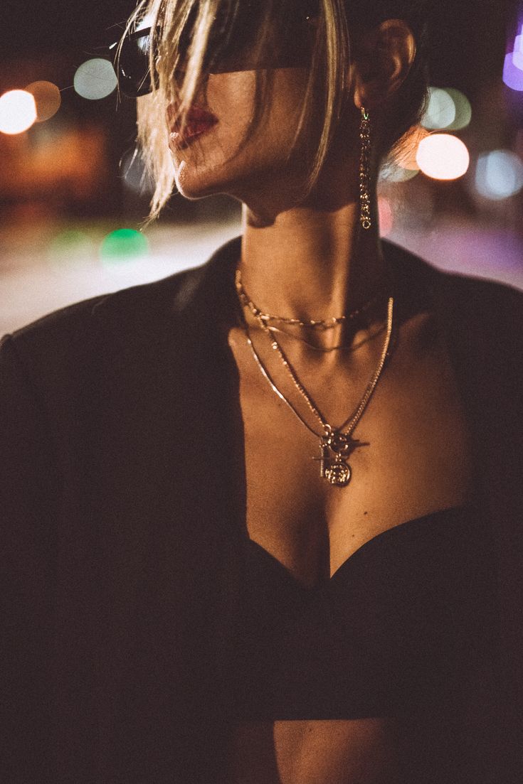 a woman wearing a black shirt and some necklaces on her neck with lights in the background