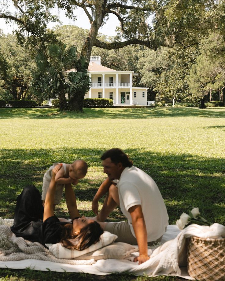 two people sitting on a blanket in front of a white house with trees and grass