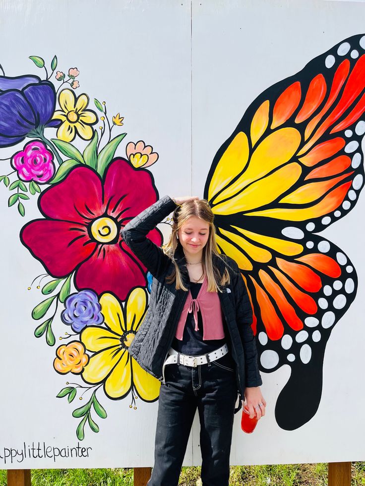 a woman standing in front of a butterfly painted on the side of a wall with flowers