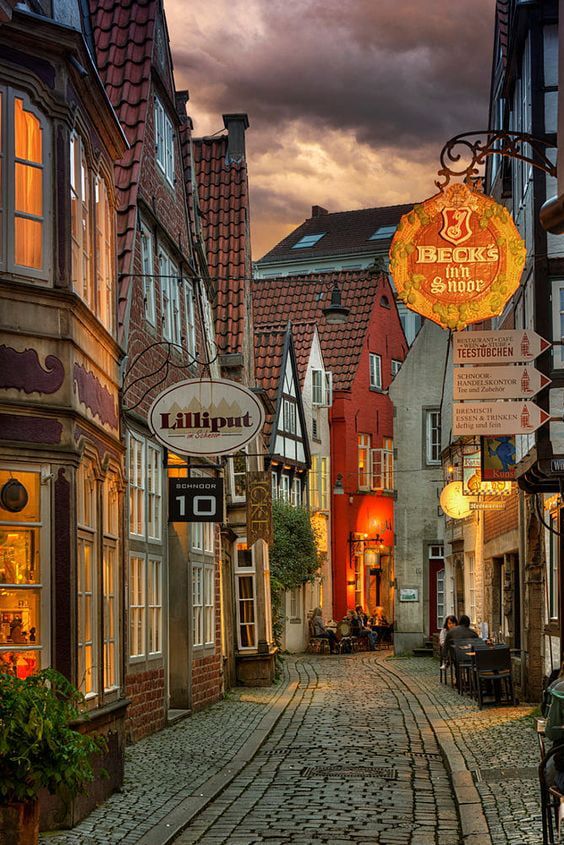 a cobblestone street lined with shops and restaurants under a cloudy sky at dusk